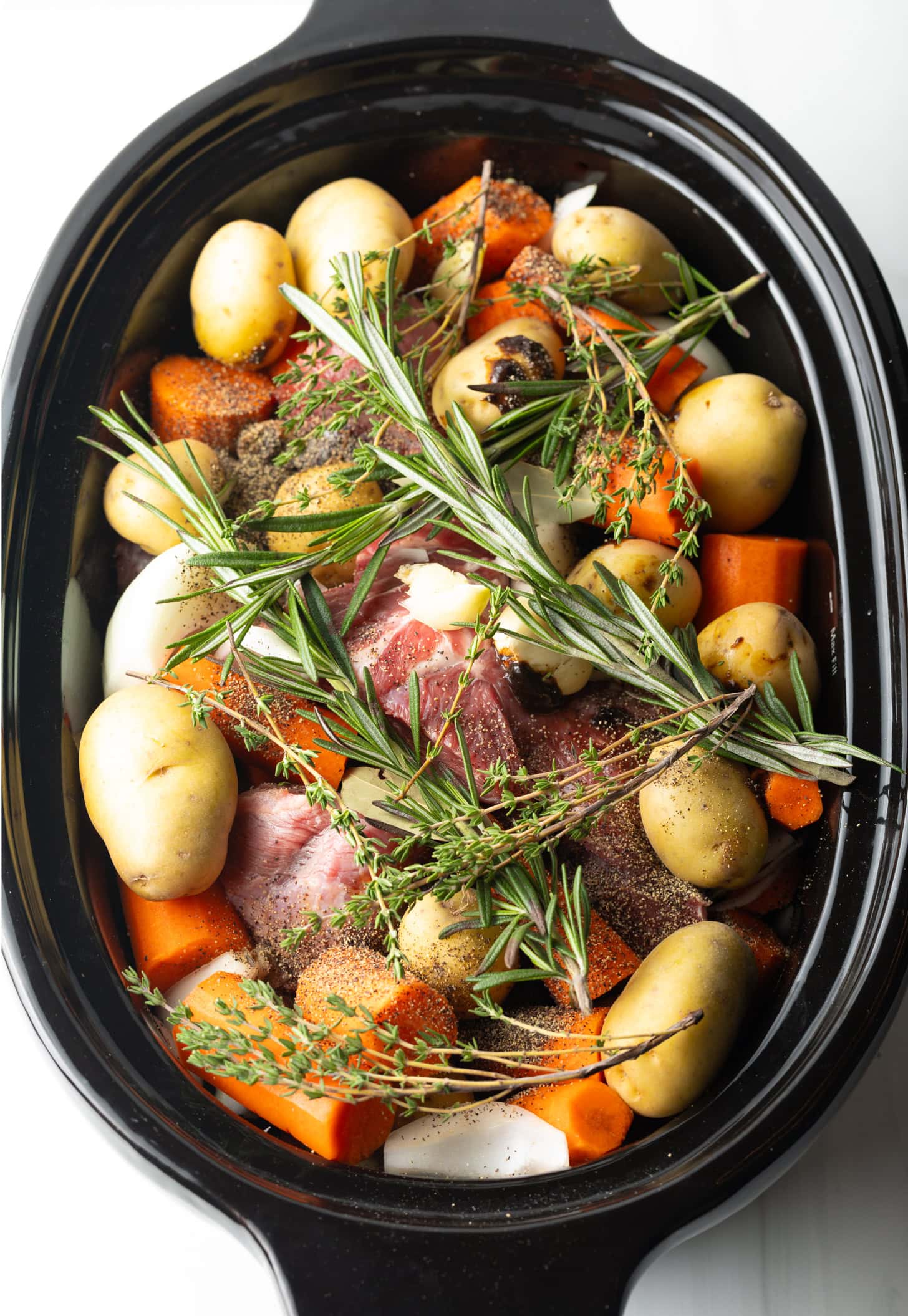 Top down in a slow cooker: Large raw beef roast is topped and surrounded by whole baby potatoes, chunks of carrots, and fresh thyme and rosemary sprigs.
