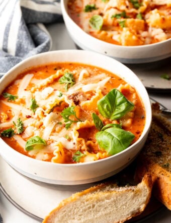 White bowl loaded with lasagna soup, garnished with two fresh basil leaves. A slice of baguette is on a white plate under the bowl.