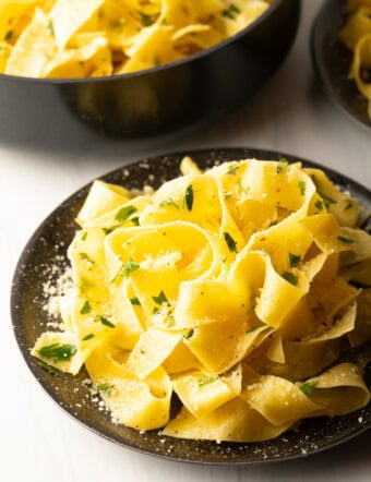Top down view of a black bowl loaded with wide pappardelle pasta topped with fresh herbs and grated parmesan cheese.