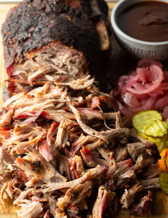 Shredded smoked pork butt on a cutting board, with ramekin of bbq sauce, stack of pickles, and pickled red onions.