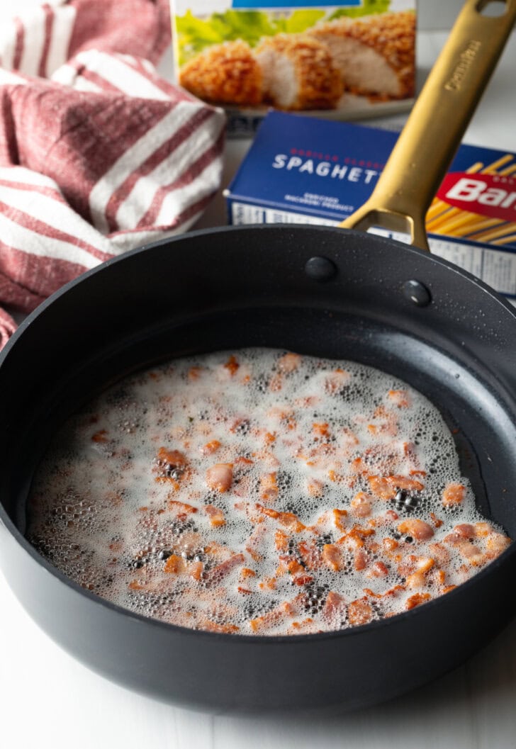 Cooking pieces of bacon in oil in a deep black skillet.