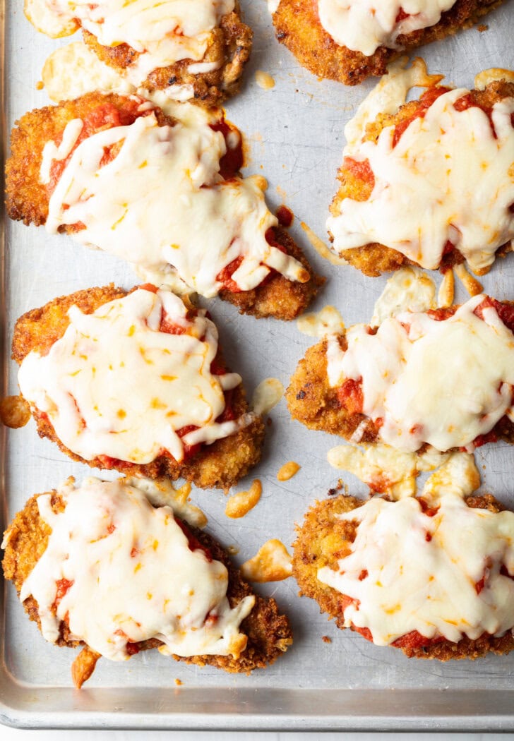 Top down view of 6 pieces of breaded baked veal on a baking sheet, topped with melted mozzarella cheese