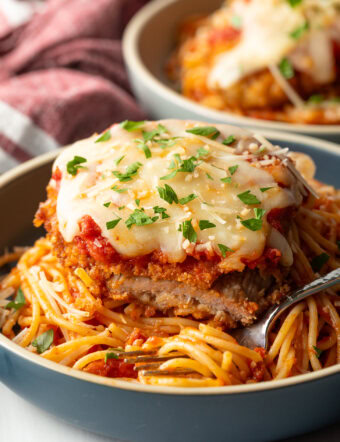 A cutlet of veal parmesan on top of spaghetti. A bite has been removed from the veal to show the juicy inside.