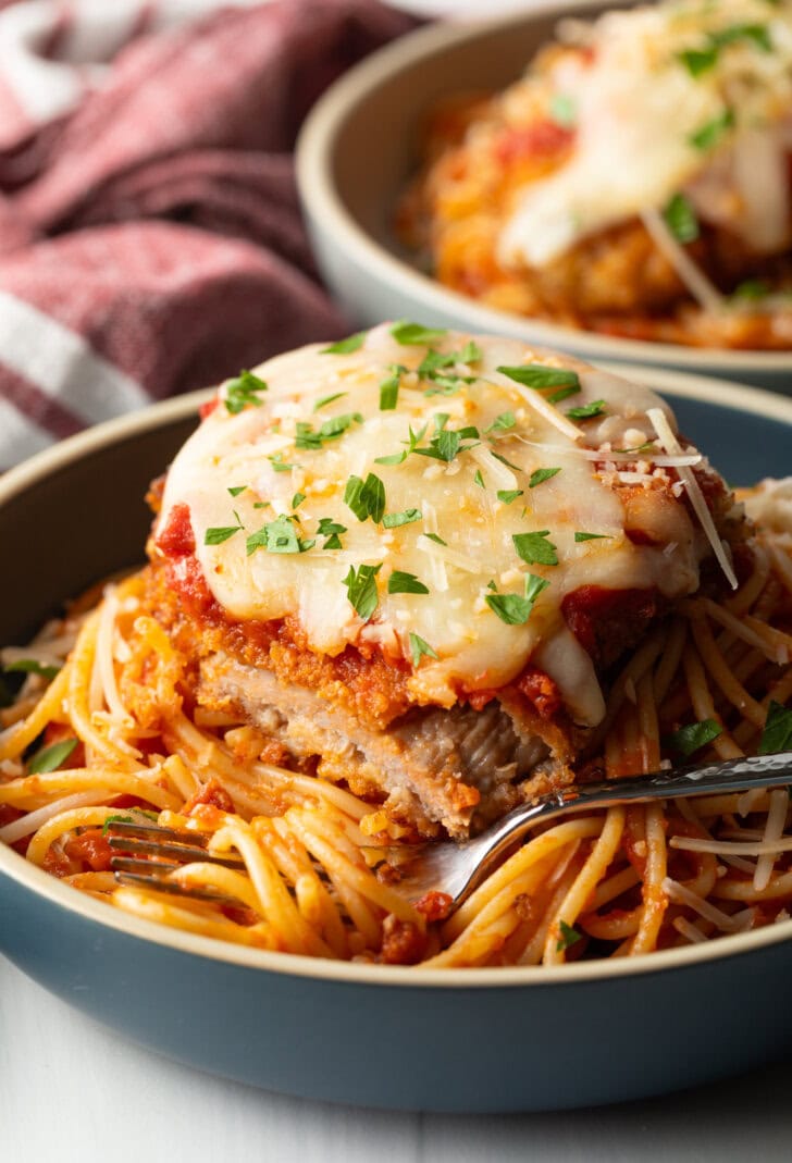 A cutlet of veal parmesan on top of spaghetti. A bite has been removed from the veal to show the juicy inside.