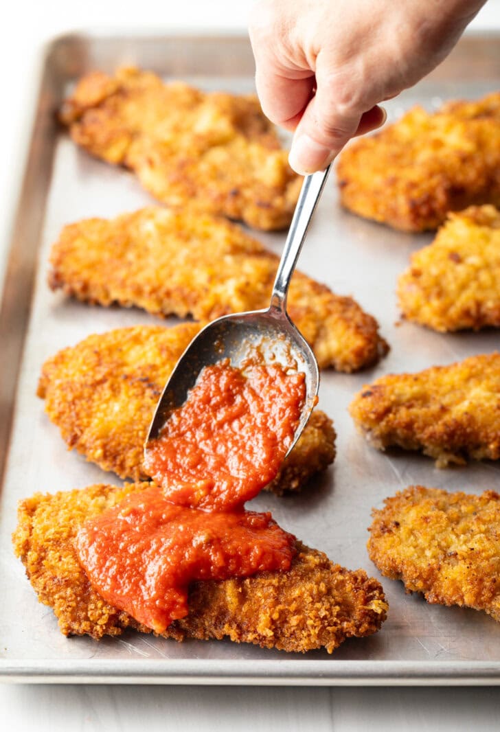 Hand with metal spoon adding marinara sauce on top of a breaded veal cutlet.