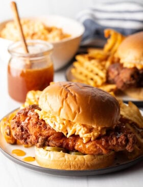Honey pepper pimento chicken sandwich on a plate. In the background is a jar of honey, and second sandwich with waffle fries.