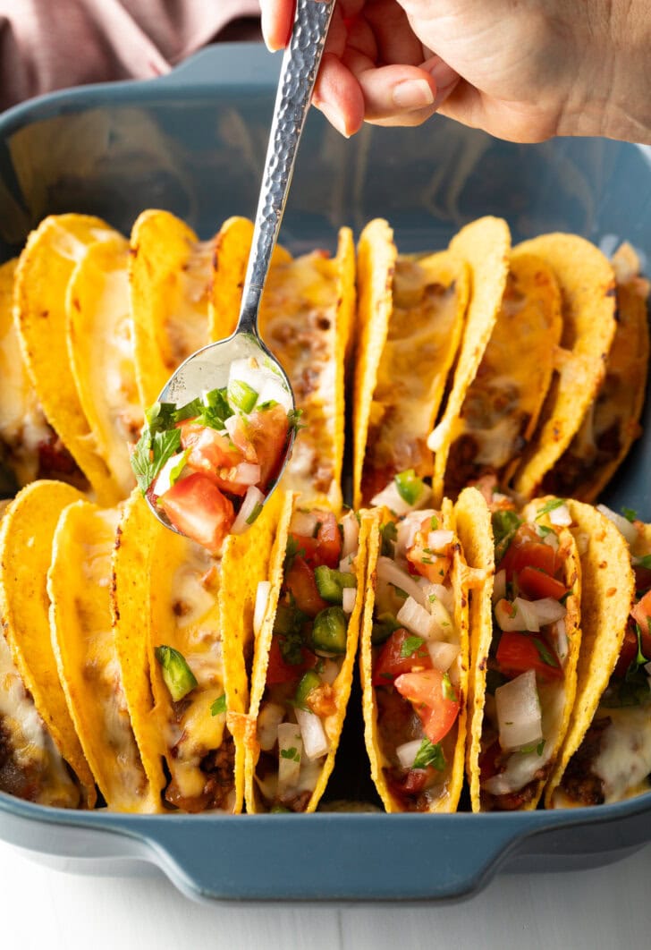 Spoon adding pico de gallo to a row of hard shell tacos.