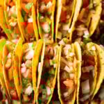 Two rows of a dozen loaded yellow corn hard shell tacos, standing upright in a large baking dish.