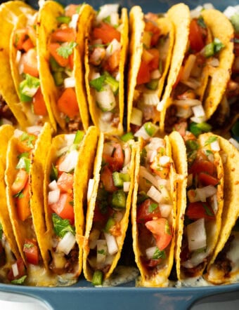 Two rows of a dozen loaded yellow corn hard shell tacos, standing upright in a large baking dish.