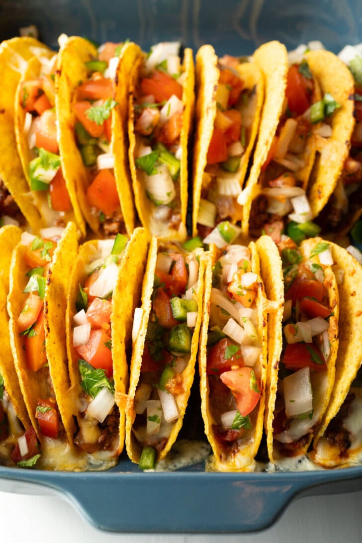 Two rows of a dozen loaded yellow corn hard shell tacos, standing upright in a large baking dish.