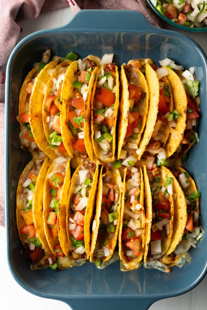 Two rows of a dozen loaded yellow corn hard shell tacos, standing upright in a large baking dish.