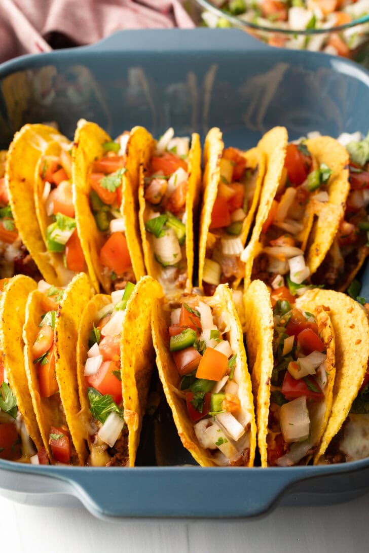 Two rows of a dozen loaded yellow corn hard shell tacos, standing upright in a large baking dish.