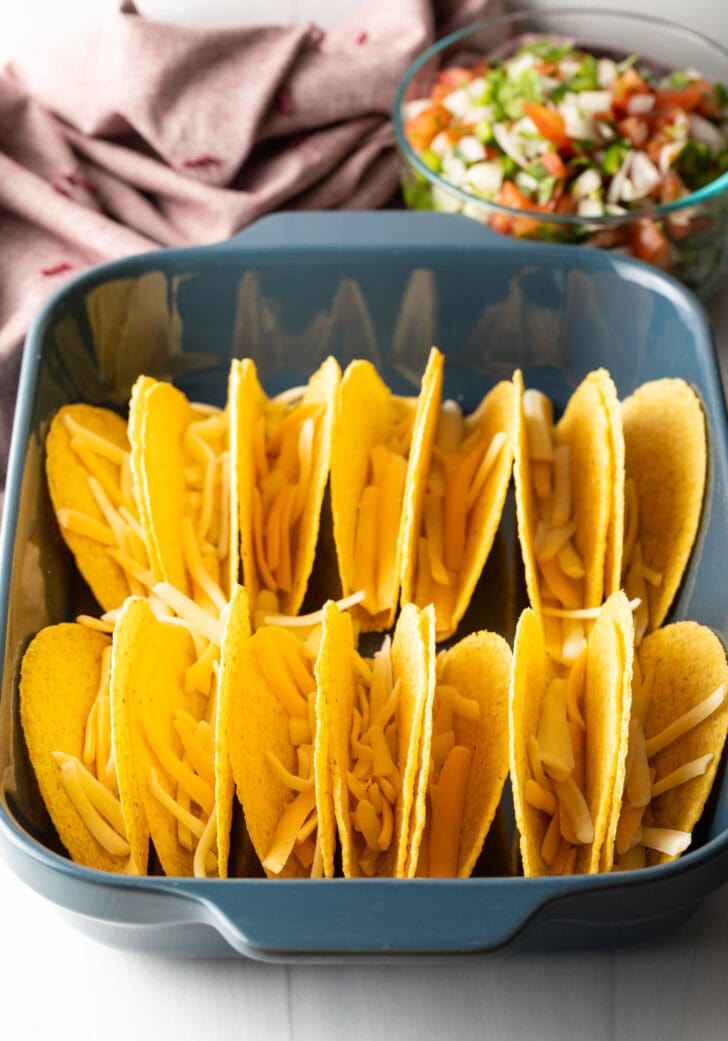Two rows of a dozen yellow corn hard shell tacos, standing upright in a large baking dish. Shredded cheese is in the tacos.