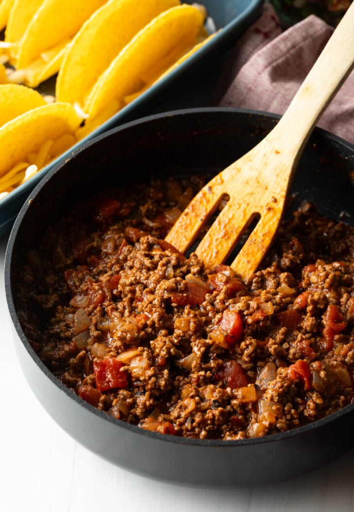 Wooden spatula stirring ground beef taco mixture in a large black deep skillet.