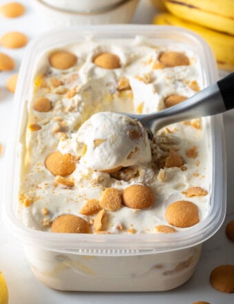 Container of no-churn banana pudding ice cream topped with nilla wafers, with a metal scooper scooping out a portion.