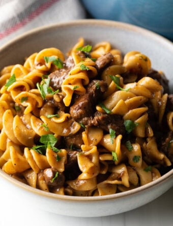 White bowl with serving of beef and noodles, topped with fresh chopped parsley.