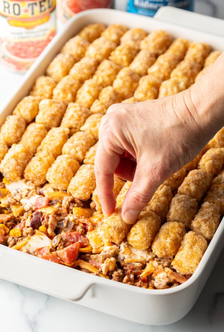 Hand adding frozen tater tots to the top of a baking dish casserole.