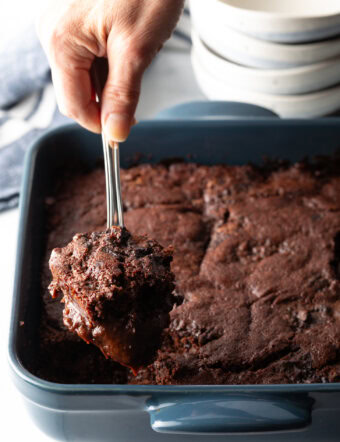 Hand using a metal spoon to scoop a serving of pudding cake from the baking pan.