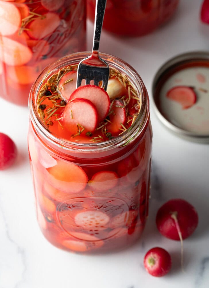 Fork picking out several pickled radishes from a large glass jar.