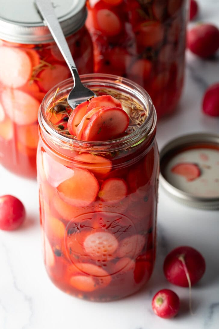 Fork picking out several pickled radishes from a large glass jar.