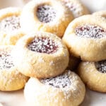 Several raspberry thumbprint cookies stacked two-high on a white plate, dusted with powdered sugar.