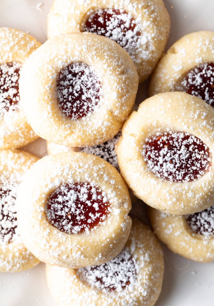 Top down view view of several raspberry thumbprints cookies stacked two-high, and three cookies in the foreground, all dusted with powdered sugar.