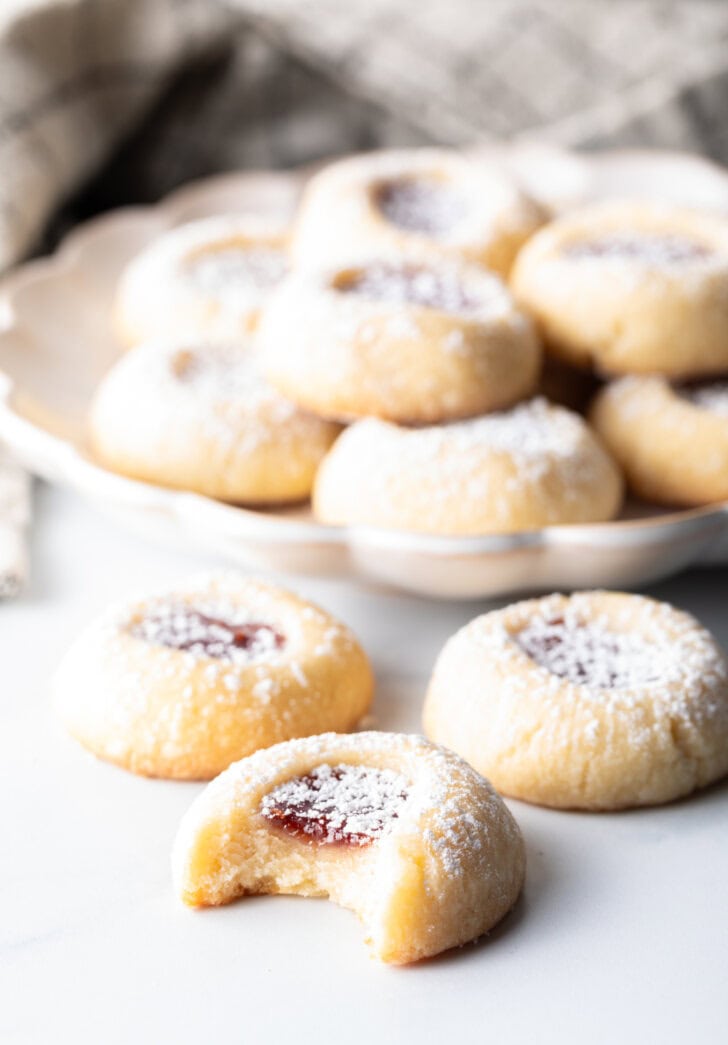 Close view of several thumbprint cookies stacked two-high, and three cookies in the foreground, all dusted with powdered sugar. A thumbprint cookie has a bite taken out.
