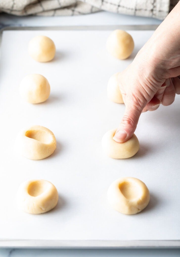 Using a thumb to press large dimples into balls of cookie dough on a baking sheet.