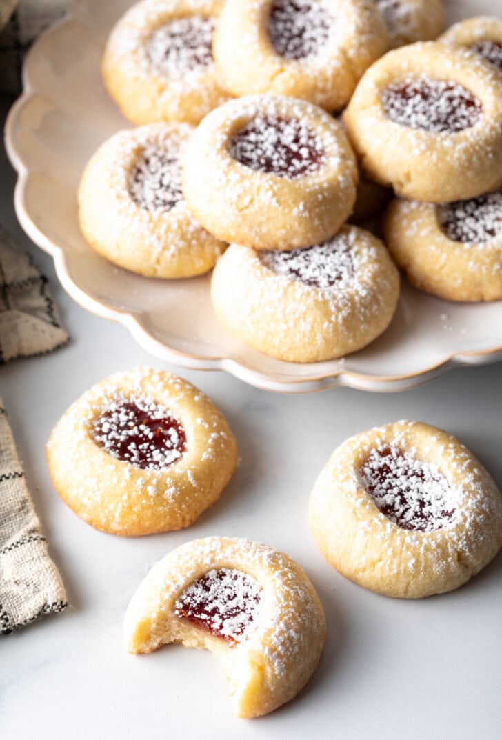 Top down view of several thumbprint raspberry cookies stacked two-high, and three cookies in the foreground, all dusted with powdered sugar.