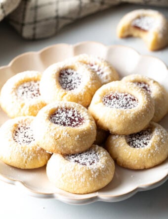 Several raspberry thumbprint cookies stacked two-high on a white plate, dusted with powdered sugar.