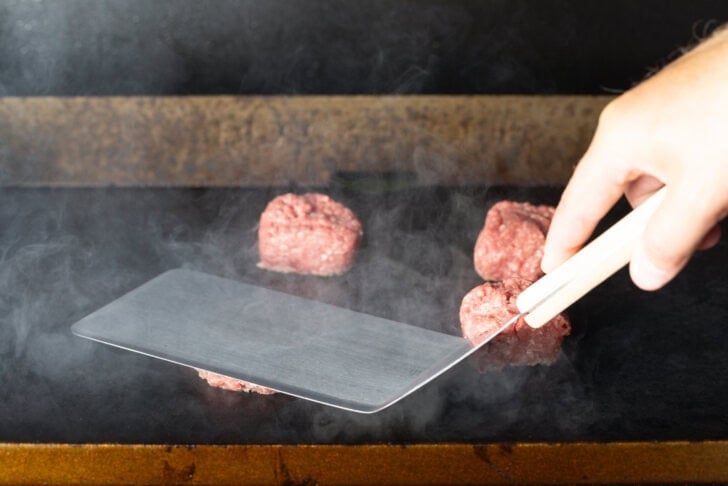 Large metal spatula smooshing a hamburger patty on a blackstone grill.