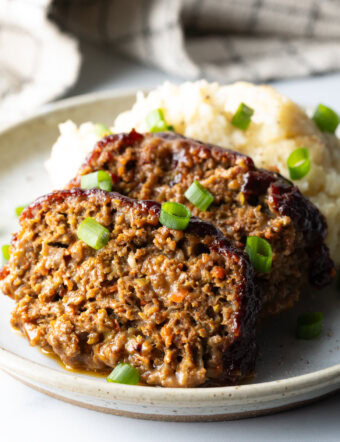2 slices of meatloaf on a plate with a scoop of mashed potatoes, all topped with chopped green onions for garnish.
