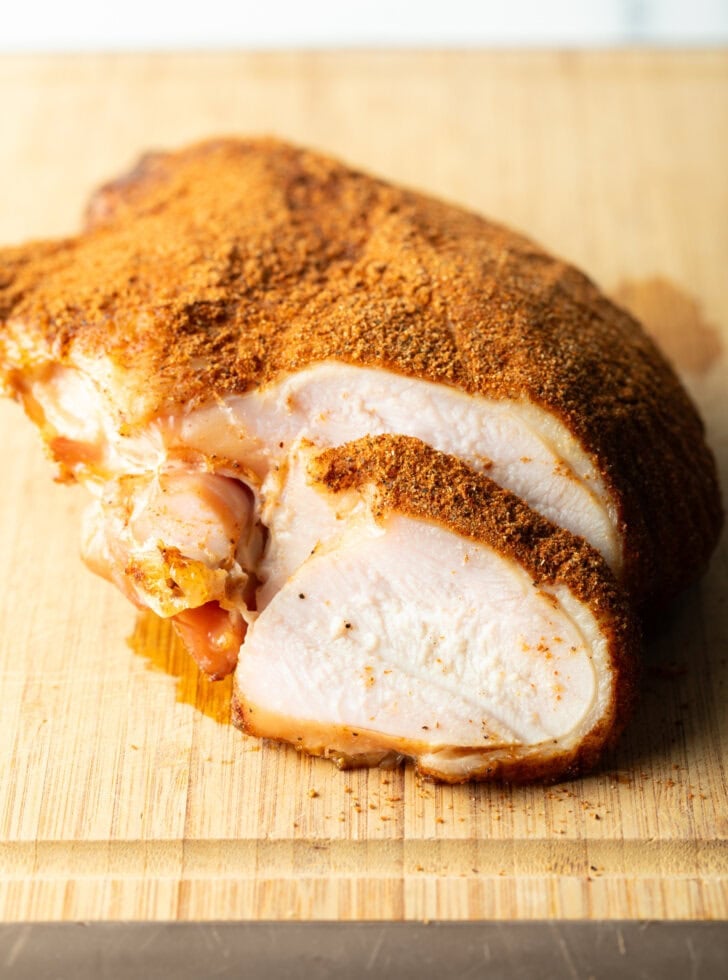 Spice-rubbed smoked turkey breast on a wood cutting board, a slice is in front of the breast.