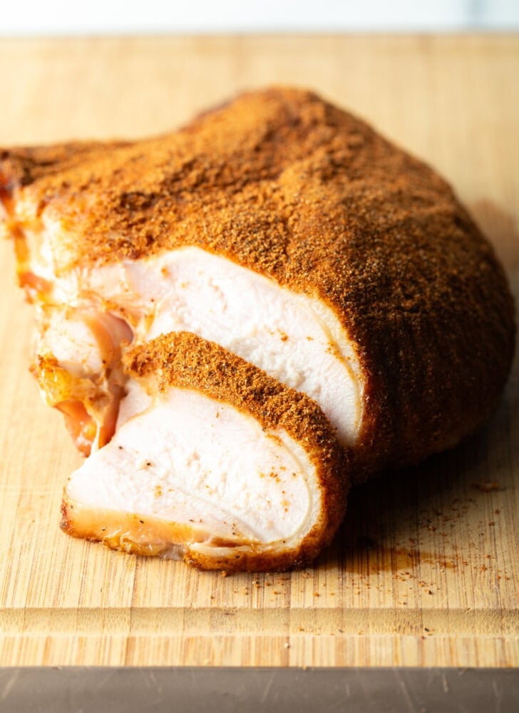 Spice-rubbed smoked turkey breast on a wood cutting board, a slice is in front of the breast.