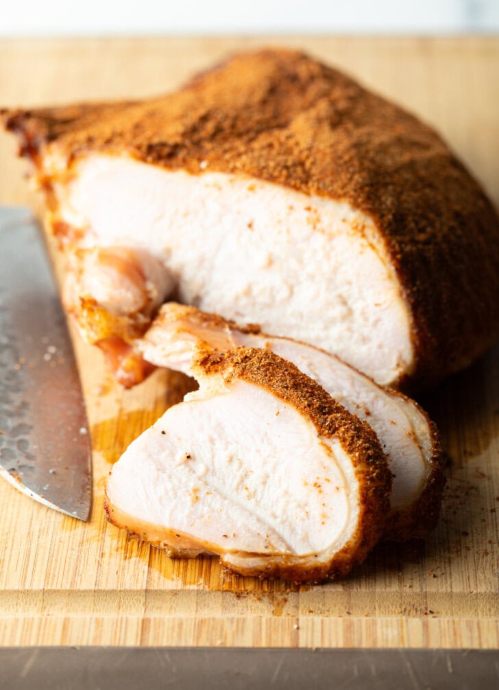 Spice-rubbed smoked turkey breast on a wood cutting board, two slices are in front of the breast.