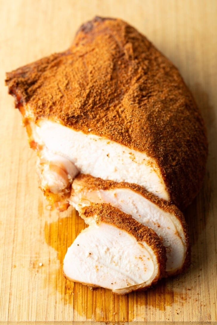 Spice-rubbed smoked turkey breast on a wood cutting board, two slices are in front of the breast.