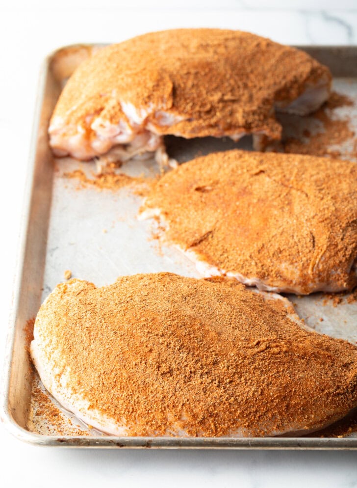 Three raw turkey breasts rubbed with seasonings, on a metal baking sheet.
