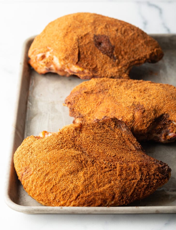 Three smoked turkey breasts rubbed with seasonings, on a metal baking sheet.