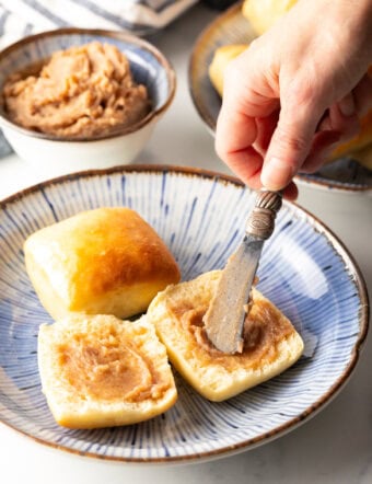 Hand using a small knife to spread cinnamon butter on both sides of an open yeast roll.