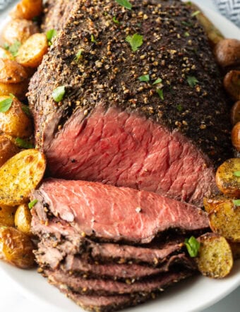 Cooked beef round roast on a metal baking pan. The front half of the roast has been thinly sliced and the meat is surrounded by roasted baby potatoes.
