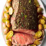 Cooked beef round roast on a metal baking pan. The front half of the roast has been thinly sliced and the meat is surrounded by roasted baby potatoes.