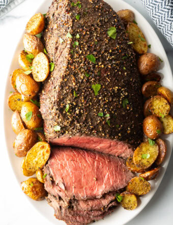 Cooked beef round roast on a metal baking pan. The front half of the roast has been thinly sliced and the meat is surrounded by roasted baby potatoes.