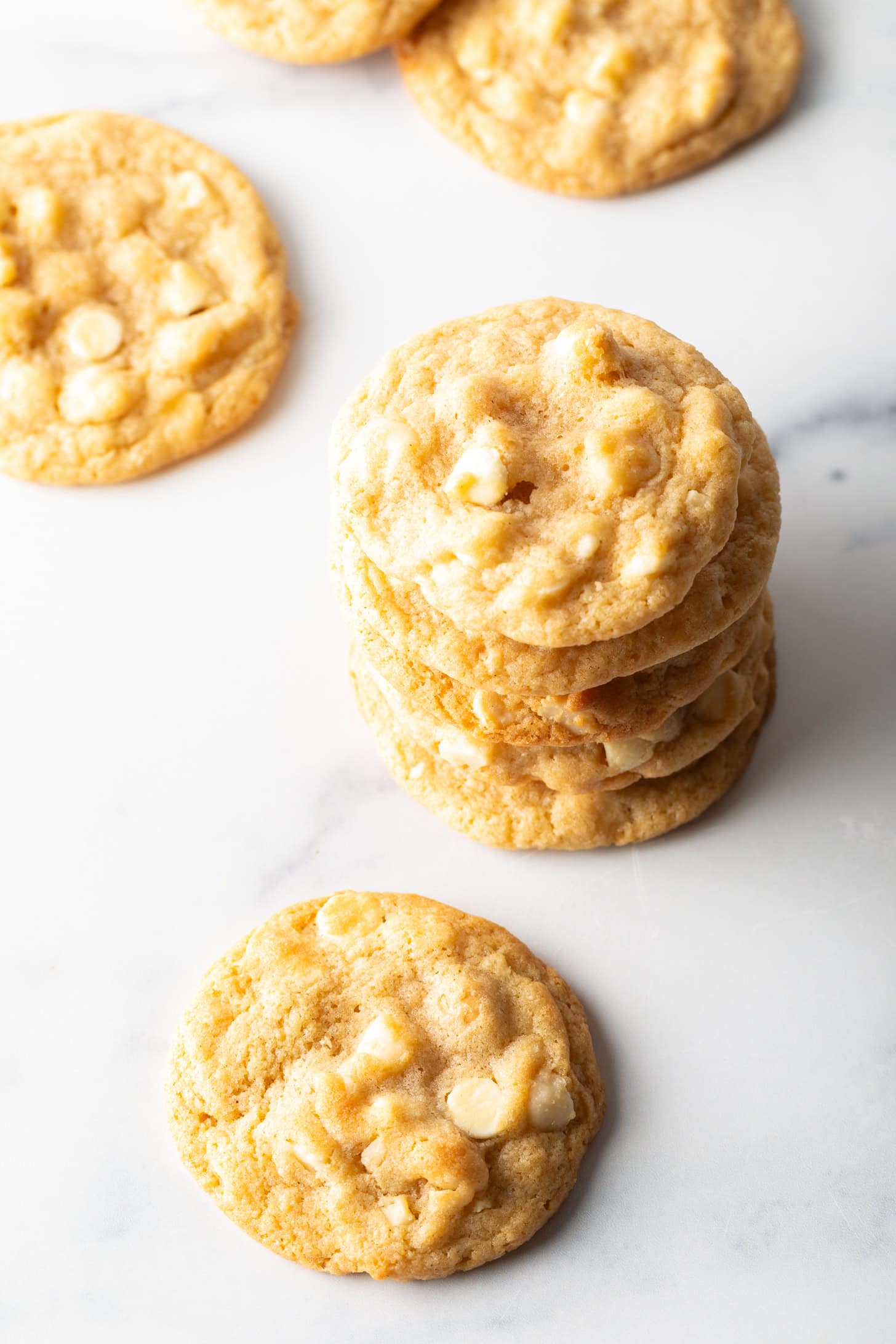 A stack of 5 macadamia nut cookies with white chocolate chips. Another cookie is laying in front of the stack.