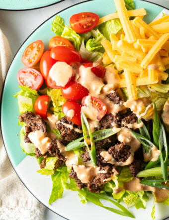 Big mac burger salad with shredded lettuce, ground beef, halved grape tomatoes, shredded cheese, pickles, and sliced green onion, all topped with a drizzle of Thousand island dressing.