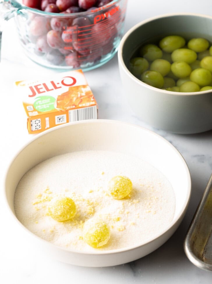 Shallow bowl with 3 green grapes being rolled in a sugar and Jello powder mixture.