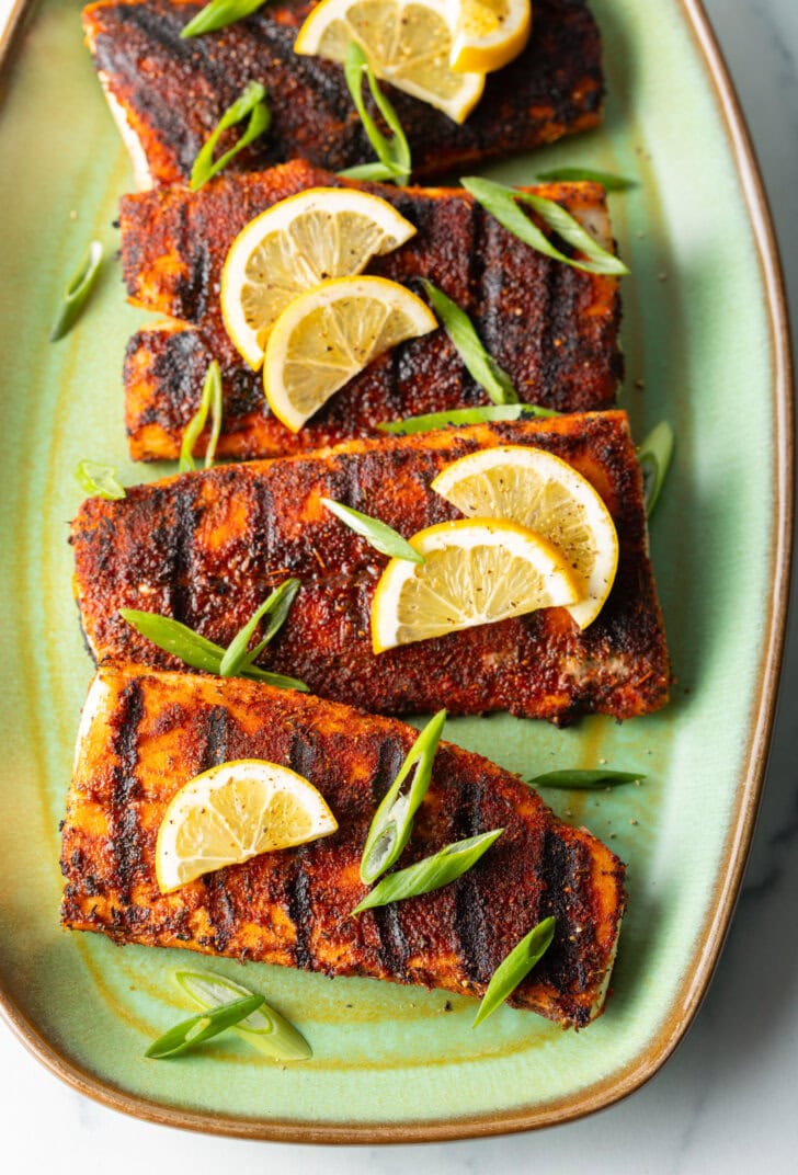 Top down view of 4 pieces of blackened and grilled fish, all topped with slices of lemon and green onions.
