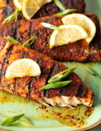 Close view of a fillet of grilled mahi mahi topped with a slice of lemon and green onions. A bite has been removed from the fish to show the white flaky inside of the meat.