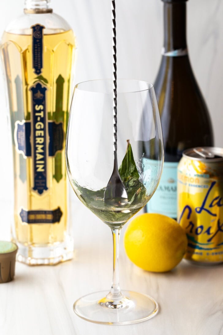 Wine glass with mint leaves being muddled with a metal spoon.