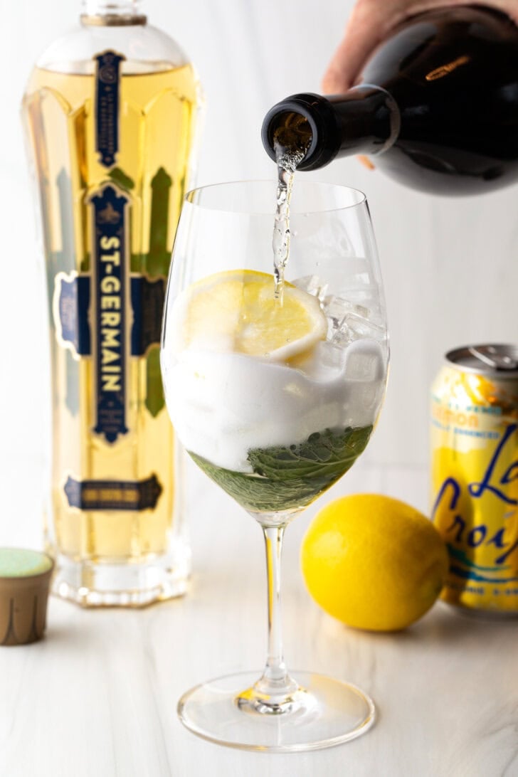 Wine glass with alcohol, mint leaves, and lemon slices. In the background are a whole lemon, st. germain liquor, and a can of la croix soda. A hand is adding prosecco from a bottle.