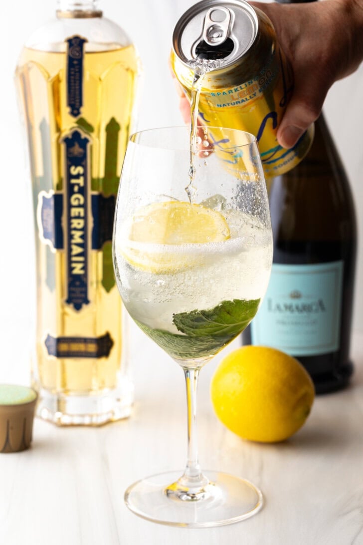 Wine glass with alcohol, mint leaves, and lemon slices. In the background are a whole lemon and bottles of champagne, st. germain liquor. A hand is adding soda from a can of la croix soda.